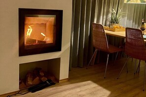 A fireplace with a fire, a dining table with chairs, and a plant on the table. - Holiday home with 1 bedroom (Julianadorp)