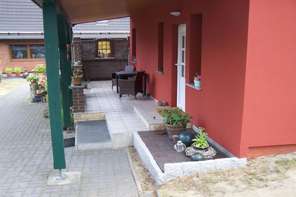 A covered outdoor area with a brick wall, a door, and a window. There are potted plants and a table with chairs.