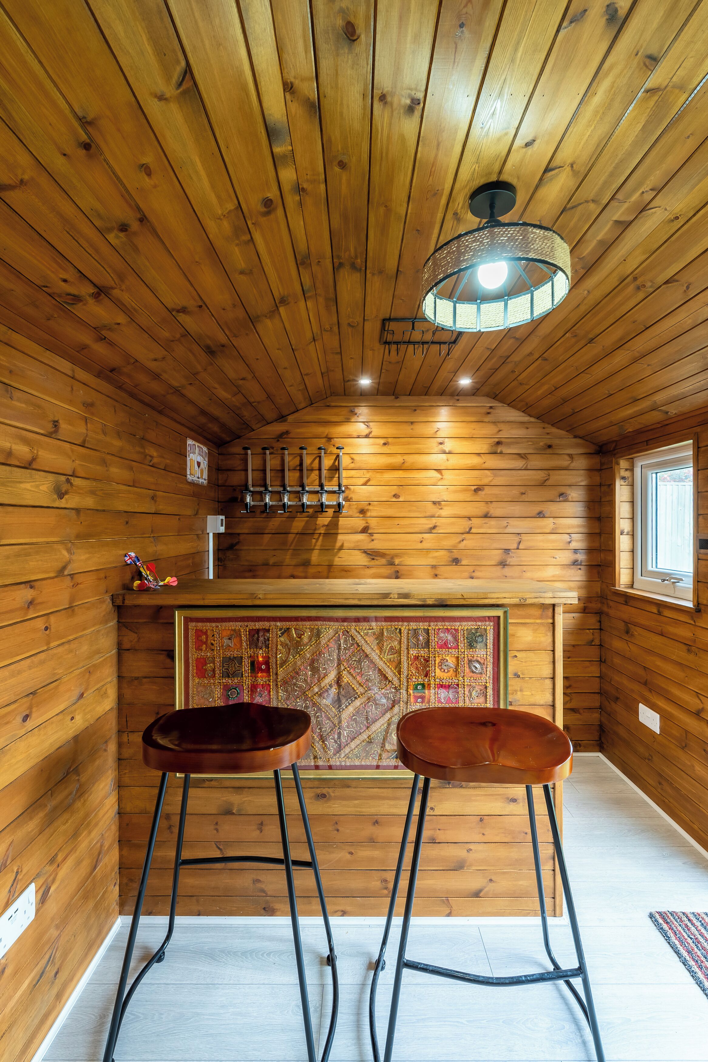 A wooden interior with a sloped ceiling, a hanging light fixture, a bar area with stools, and a decorative wall panel.