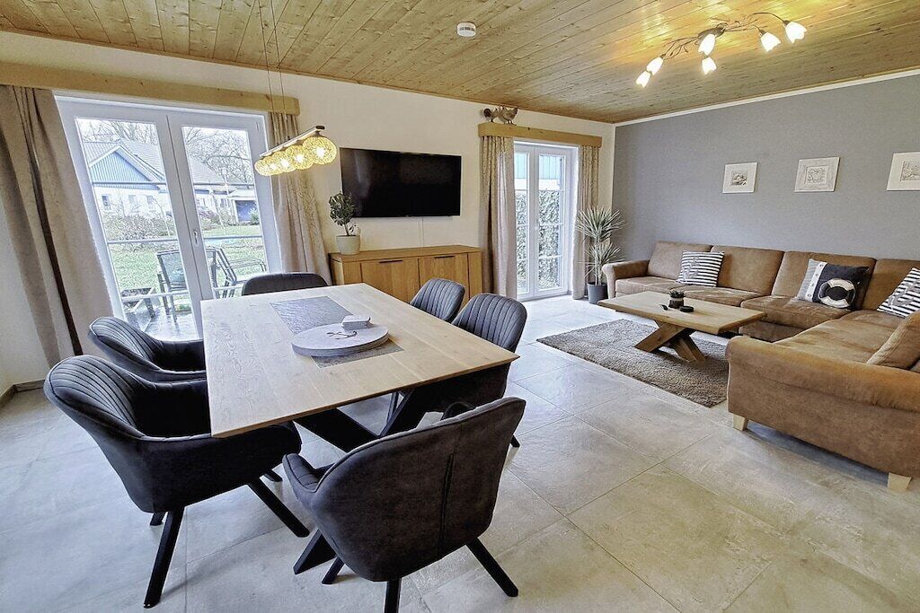 A modern living room with a dining area, a sofa set, a flat-screen TV, and a large window.
