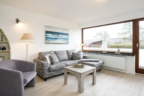 A living room with a sofa, a coffee table, a lamp, and a window with a view. - Holiday apartment with 1 bedroom (Westerland\/ Sylt)