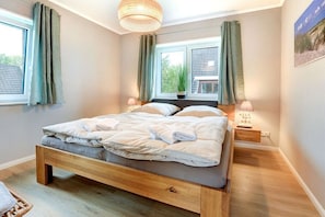 A bedroom with a wooden bed, two bedside tables with lamps, a window with curtains, and a view of greenery outside. - Holiday apartment with 2 bedrooms (St. Peter-Ording)