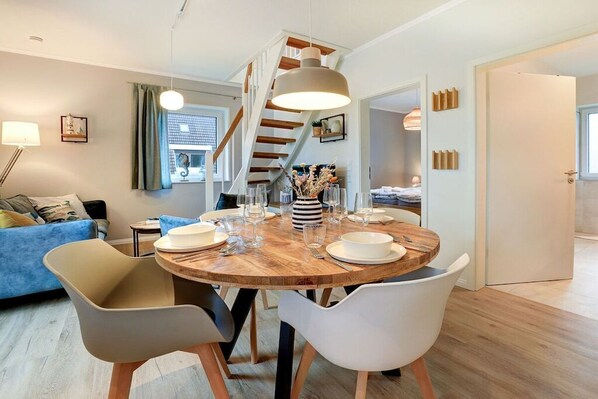 A modern dining area with a round wooden table, white chairs, and a hanging pendant light. - Holiday apartment with 2 bedrooms (St. Peter-Ording)