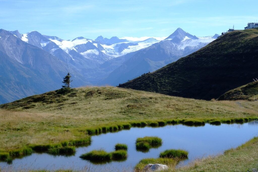 Een bergachtig landschap met een klein vijvertje, één boom en verafgelegen, besneeuwde bergtoppen.
