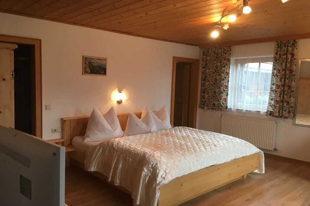 Une chambre à coucher avec un lit en bois, une table de chevet, une lampe fixée au mur, une fenêtre avec des rideaux et un radiateur.