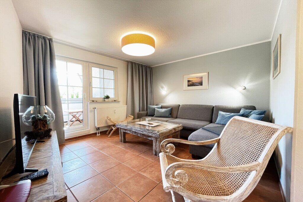 Ein Wohnzimmer mit einer Couch, einem Couchtisch, einem Korbstuhl, einem Fernseher und einem Fenster mit Vorhängen.