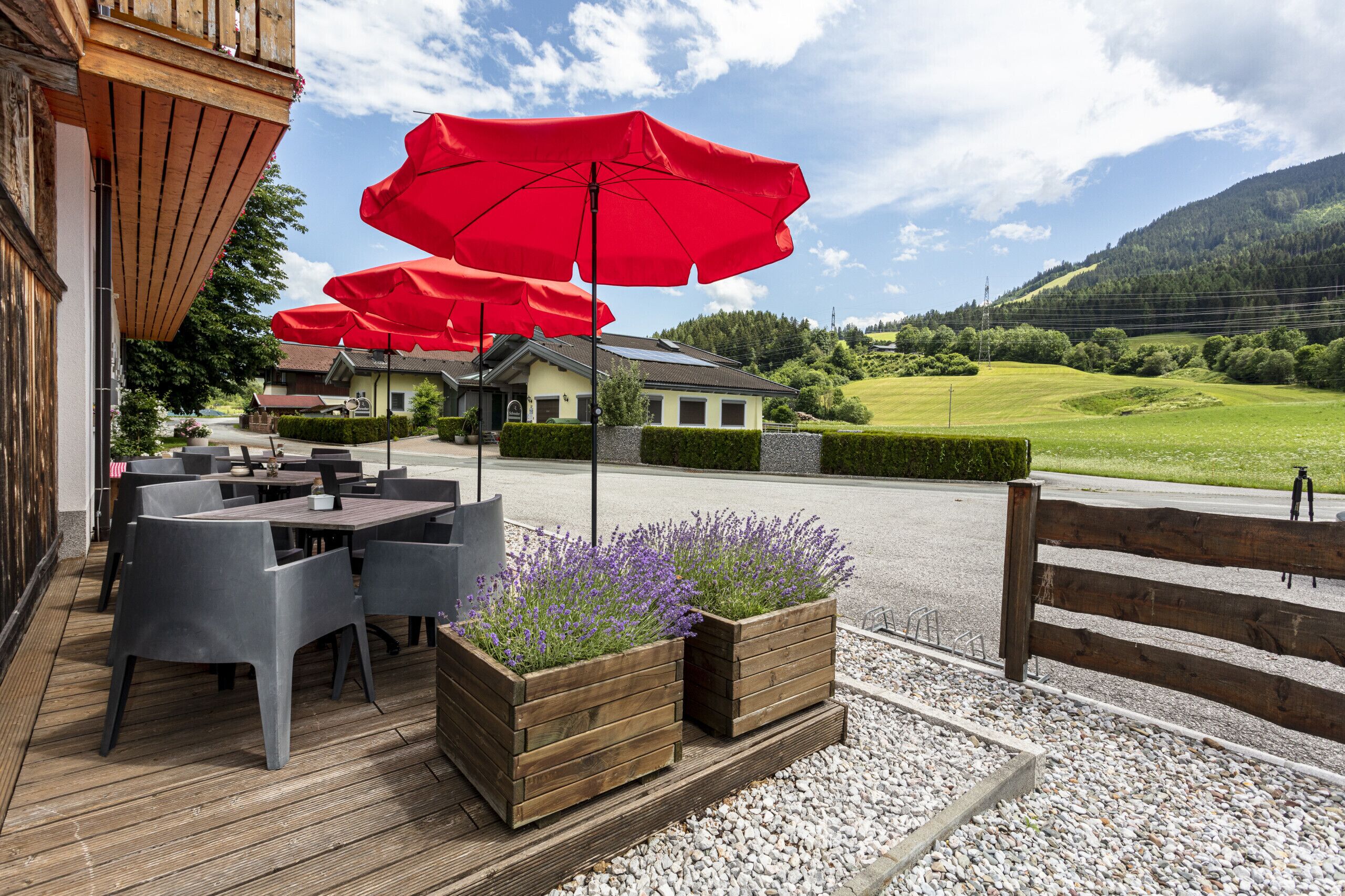 An outdoor seating area with tables and chairs, red umbrellas, and a view of a mountain landscape.