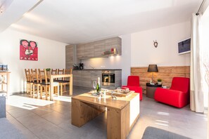 Sala de estar moderna com mesa de centro de madeira, cadeiras vermelhas, área de cozinha com armários cinzas e uma televisão na parede.