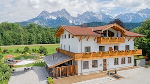 A two-story chalet with a balcony, surrounded by a garden and a pond, with mountains in the background. - Gästehaus Osterthor - Ferienwohnung Asitz, Balkon & Bergblick (Saalfelden am Steinernen Meer)