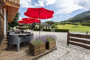 Ein Sitzbereich im Freien mit Tischen und Stühlen, roten Sonnenschirmen und Blick auf eine Berglandschaft.
