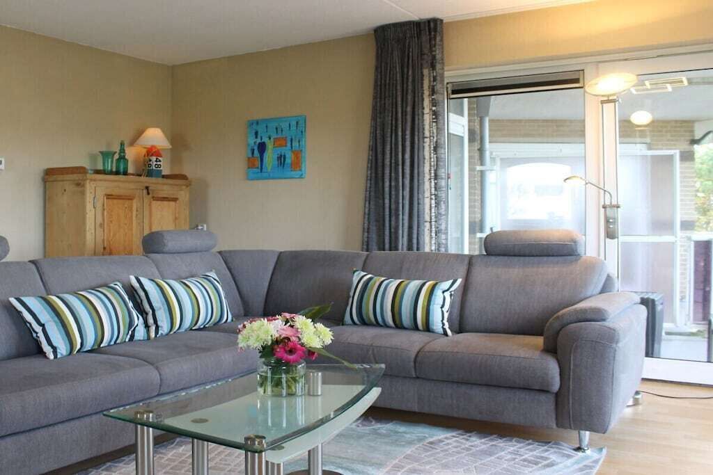 Una sala de estar con un sofá gris, cojines a rayas, una mesa de centro de vidrio con flores, un mueble de madera y un cuadro en la pared.