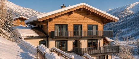 A wooden chalet with a balcony, set against a snowy mountain backdrop.