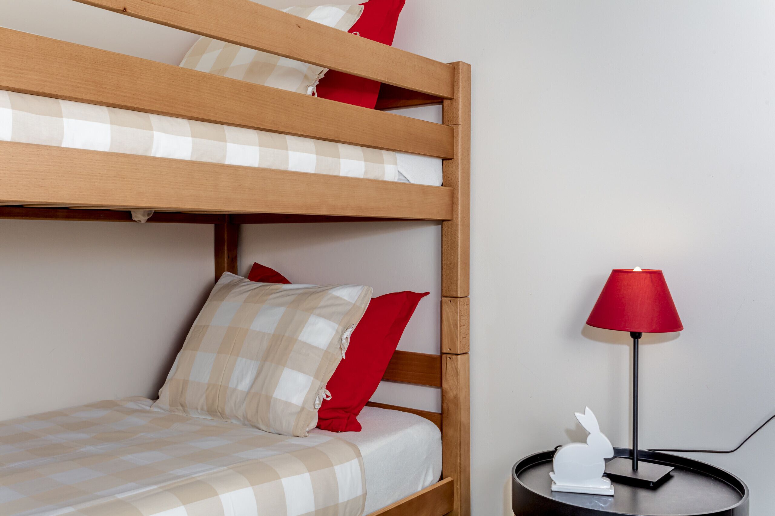 Uma cama beliche com colcha xadrez e almofadas vermelhas, uma mesa de cabeceira com abajur vermelho e uma decoração de coelho branco.