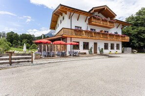 Ein zweistöckiges Gebäude mit Balkon, Außenbereich mit roten Sonnenschirmen und Bergblick im Hintergrund.