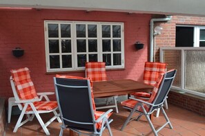Eine Terrasse mit einem Holztisch und vier Stühlen, ein Fenster mit Jalousien und eine Ziegelwand.