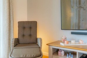 Habitació amb una cadira tapissada de gris, una taula auxiliar de fusta amb espelmes i una estanteria amb pots.
