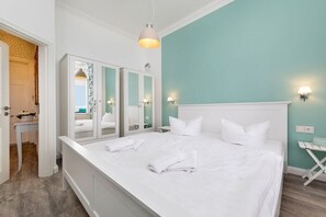 Ein ordentlich bezogenes Bett mit weißen Leinen und Kissen, ein weißer Kleiderschrank mit Spiegelüren und eine türkisblaue Wand.