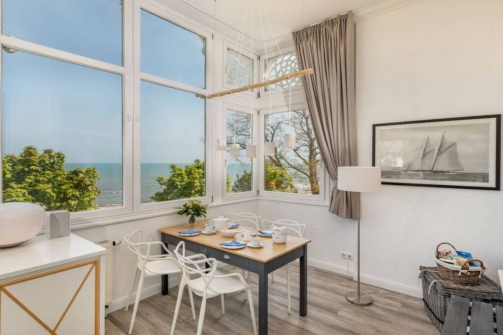 Una habitación con una mesa de comedor preparada para cuatro personas, ventanas amplias con vista al mar, una lámpara de pie y un cuadro en la pared.