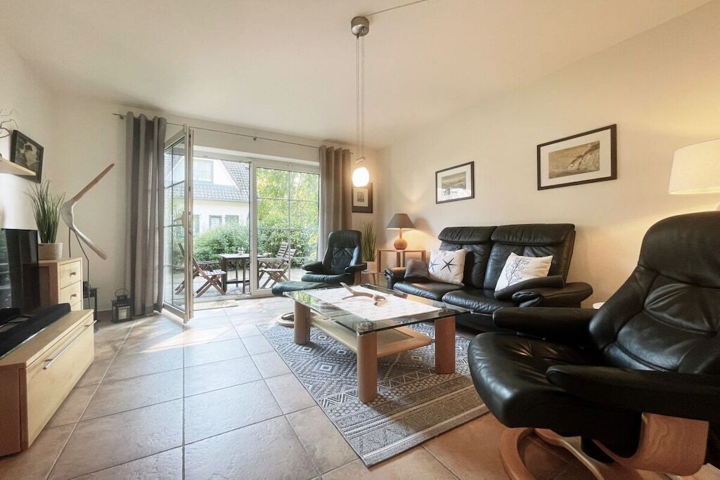 A living room with a sofa, armchairs, a coffee table, and a TV. There is a sliding glass door leading to an outdoor area with a table and chairs.