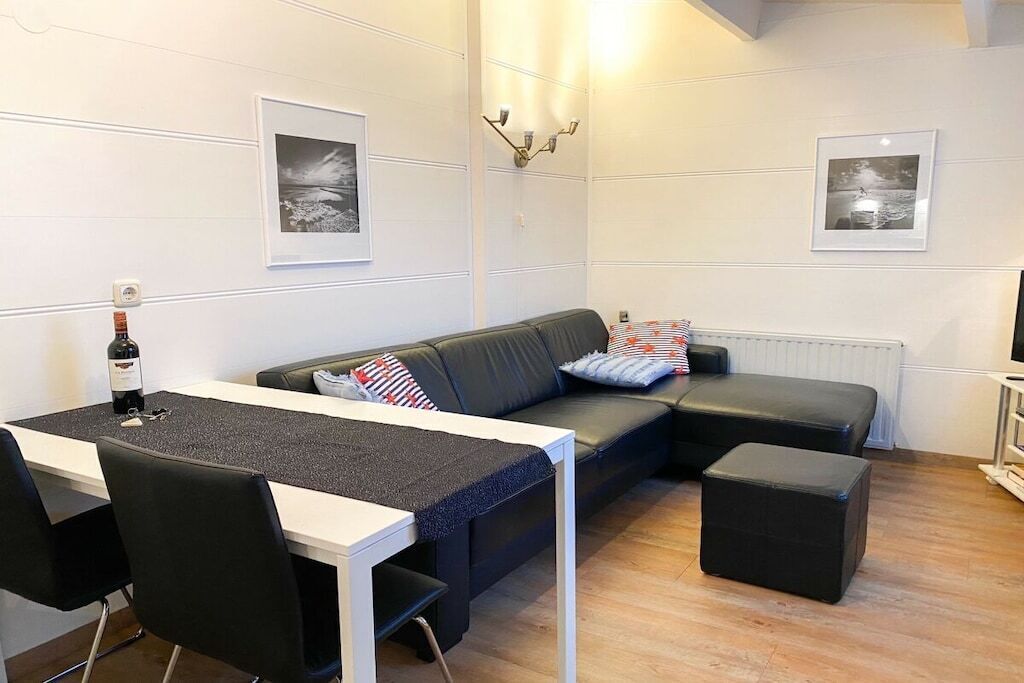 Una sala de estar moderna con un sofá, una mesa de comedor y un reposapiés negro. Hay cuadros enmarcados en la pared y una botella de vino sobre la mesa.