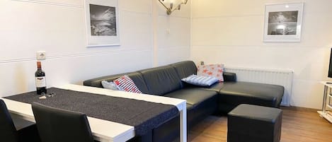 Una sala de estar moderna con un sofá, una mesa de comedor y un reposapiés negro. Hay cuadros enmarcados en la pared y una botella de vino sobre la mesa.
