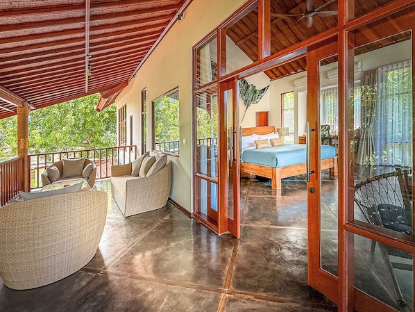 A covered outdoor seating area with wicker furniture, a bedroom with a bed and a nightstand, and a large sliding glass door leading to another room.