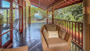 A wicker lounge chair with a cushion labeled 'Sea Esta' on a balcony with glass railings, overlooking a pool and tropical vegetation.