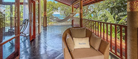 A wicker lounge chair with a cushion labeled 'Sea Esta' on a balcony with glass railings, overlooking a pool and tropical vegetation.