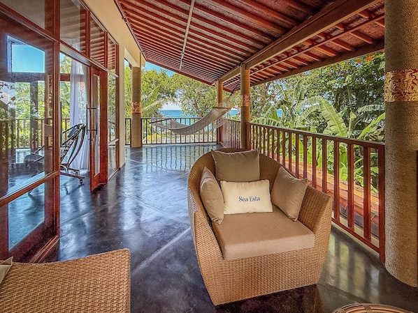 A wicker lounge chair with a cushion labeled 'Sea Esta' on a balcony with glass railings, overlooking a pool and tropical vegetation.
