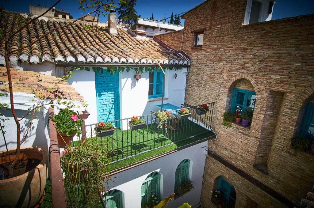 Une cour avec un bâtiment blanc aux volets bleus, un bâtiment en briques doté de fenêtres en arc de cercle et des plantes en pot.