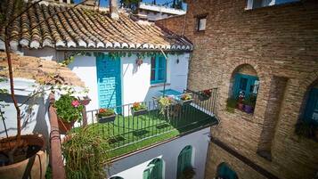 Une cour avec un bâtiment blanc aux volets bleus, un bâtiment en briques doté de fenêtres en arc de cercle et des plantes en pot.