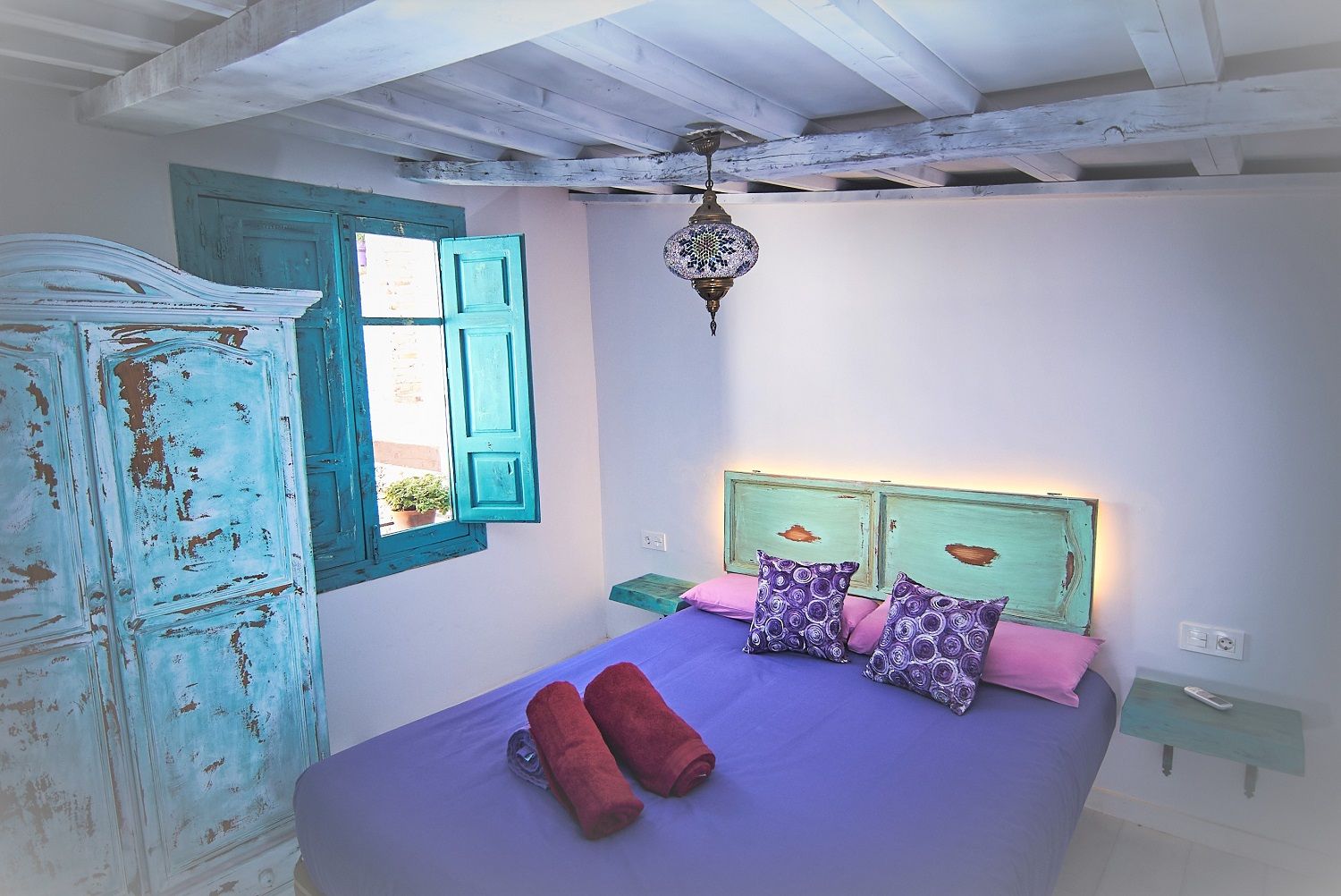 Un dormitorio con cama, armario, ventana con contraventanas azules y una lámpara colgante.