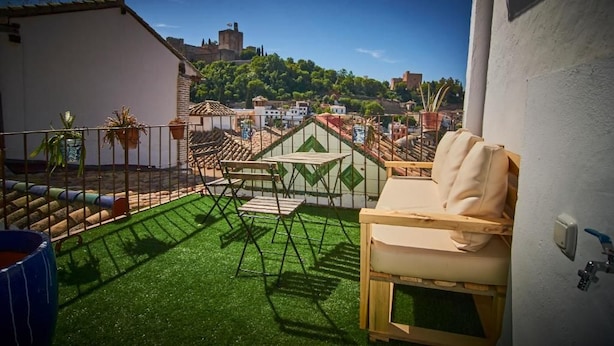 Een balkon met kunstgras, een beige bank en uitzicht op een stadsbeeld met gebouwen en een heuvel.