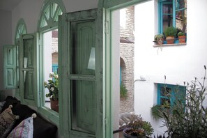 Una habitación con pared blanca, puerta verde y ventana con contraventanas azules. Hay plantas en macetas en el alféizar y una pequeña mesa con una planta.