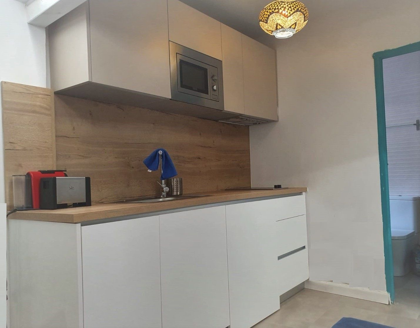 Una cocina con armarios blancos, un microondas, un lavamanos con una toalla azul y una lámpara decorativa.