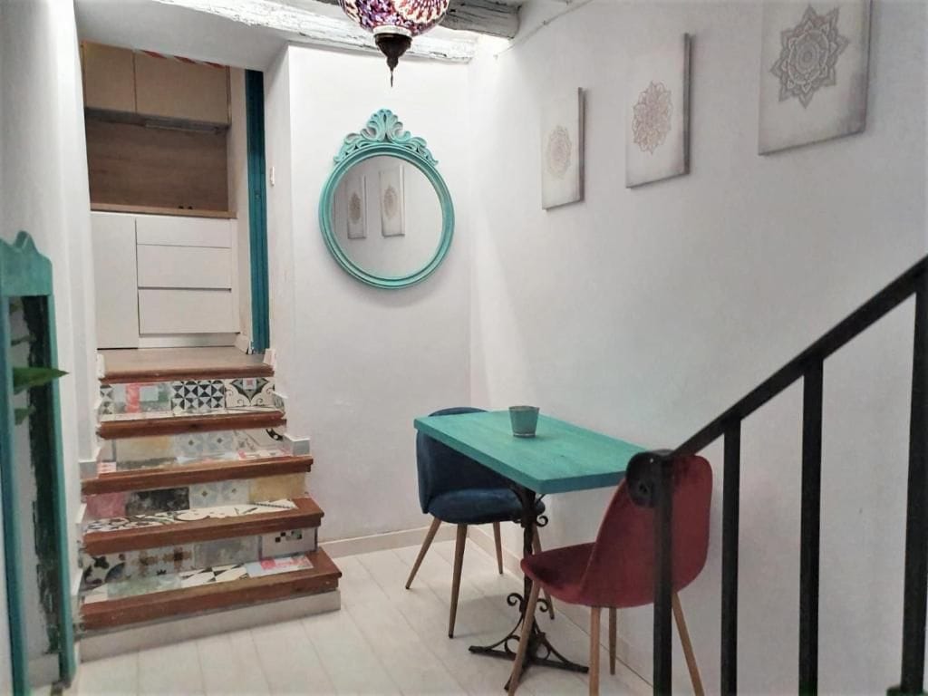 Una escalera con baldosas estampadas, una mesa color turquesa con dos sillas y un espejo decorativo en la pared.