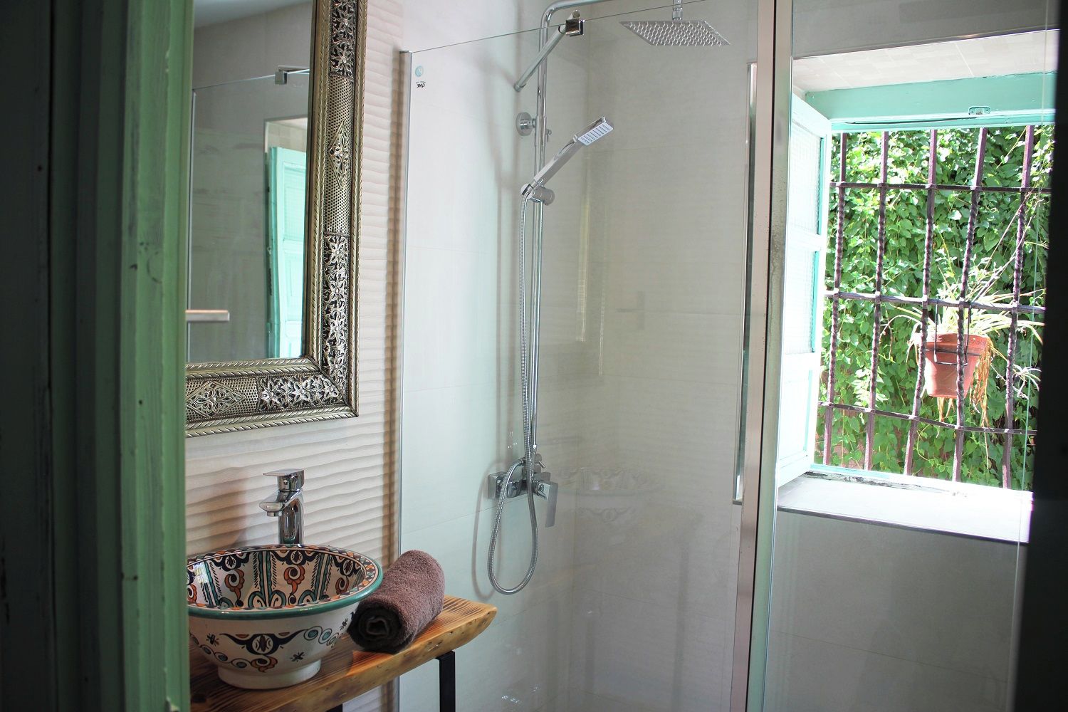 A bathroom with a shower, a sink with a decorative bowl, a mirror with an ornate frame, and a window with a view of greenery.