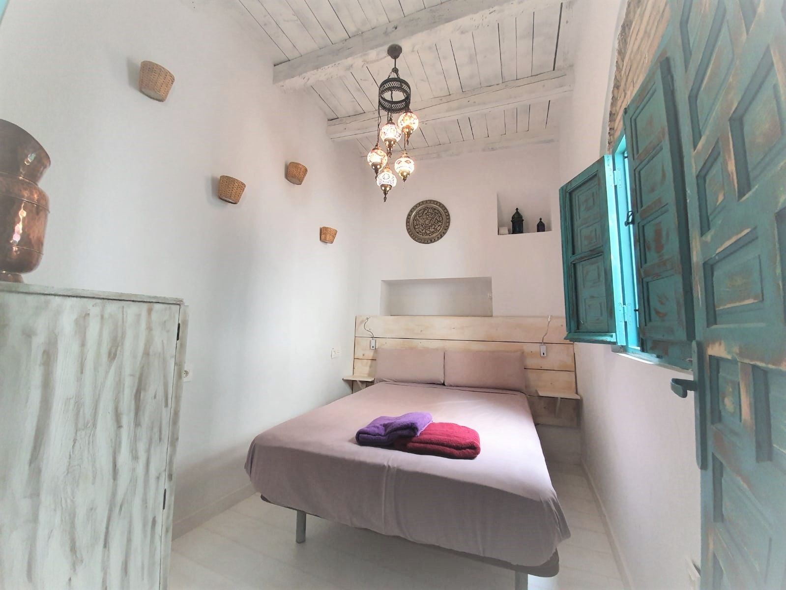 A bedroom with a bed, a wooden headboard, a hanging light fixture, and a window with shutters.