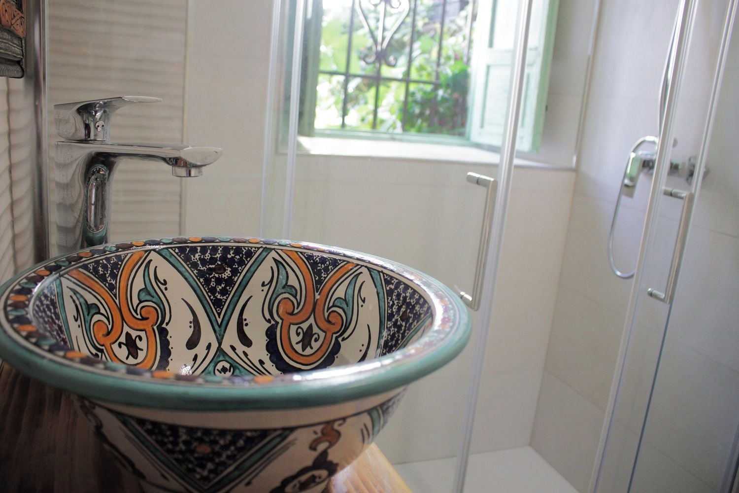 A bathroom with a decorative ceramic sink, a shower, and a window with a view of greenery.