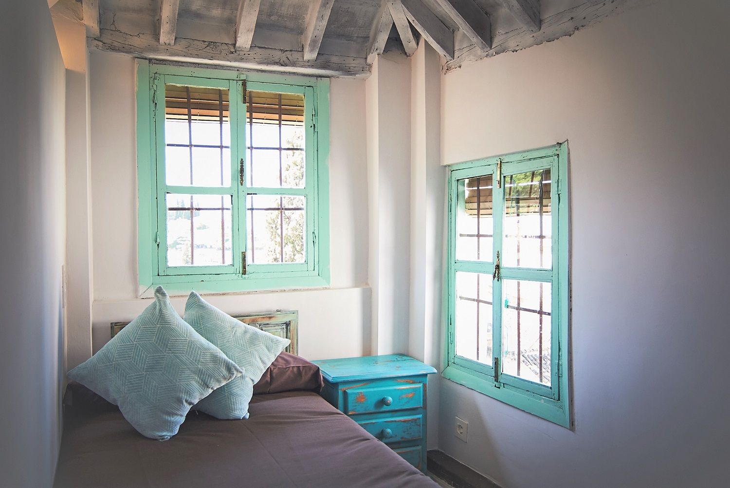 Un dormitorio con cama, dos almohadas, una mesita de noche y dos ventanas con contraventanas.