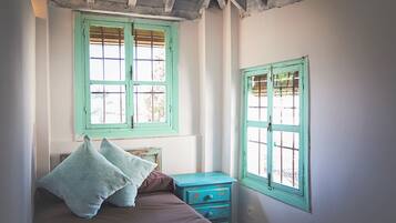 Un dormitorio con cama, dos almohadas, una mesita de noche y dos ventanas con contraventanas.