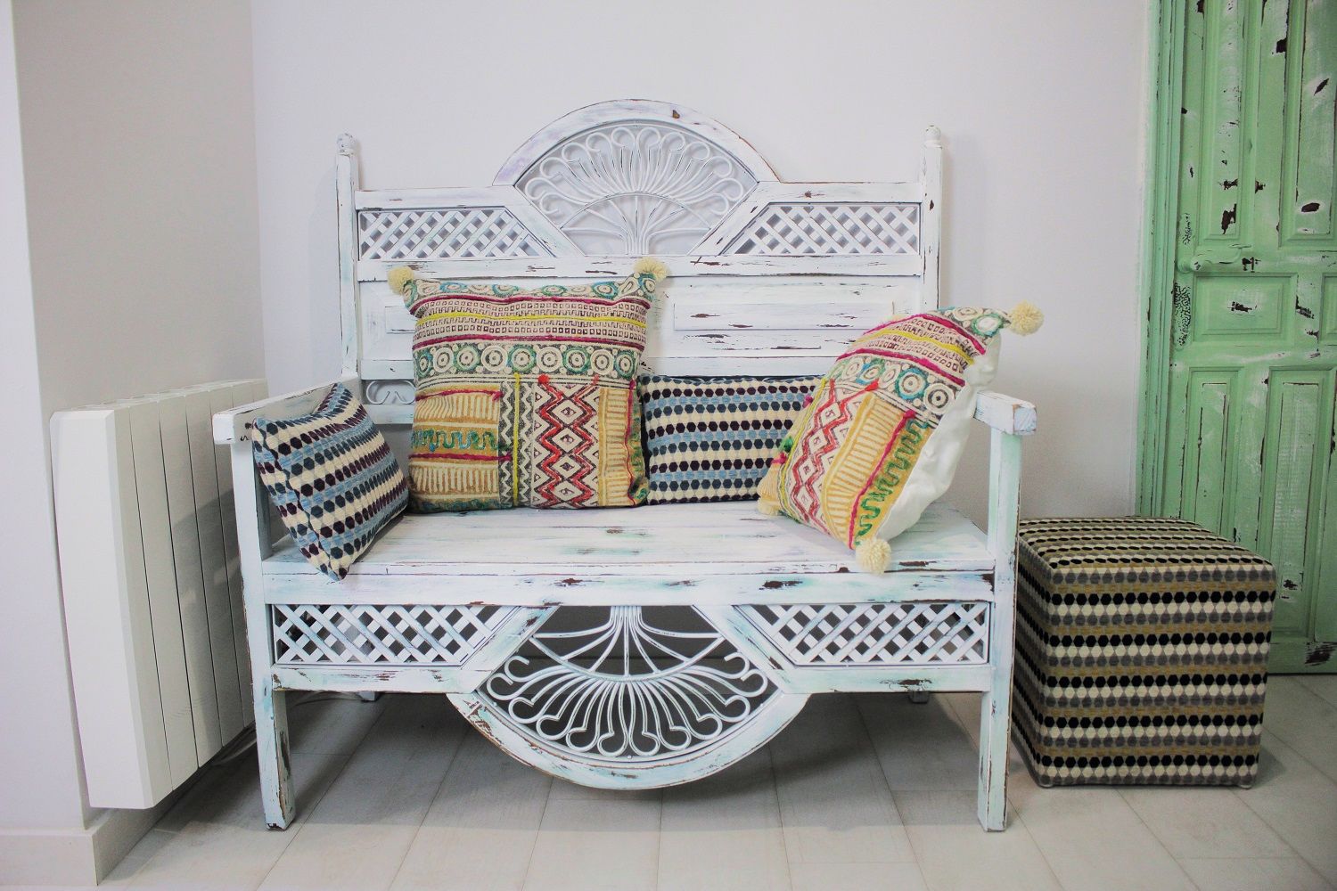 A white, distressed wooden bench with intricate carvings and multiple colorful cushions.