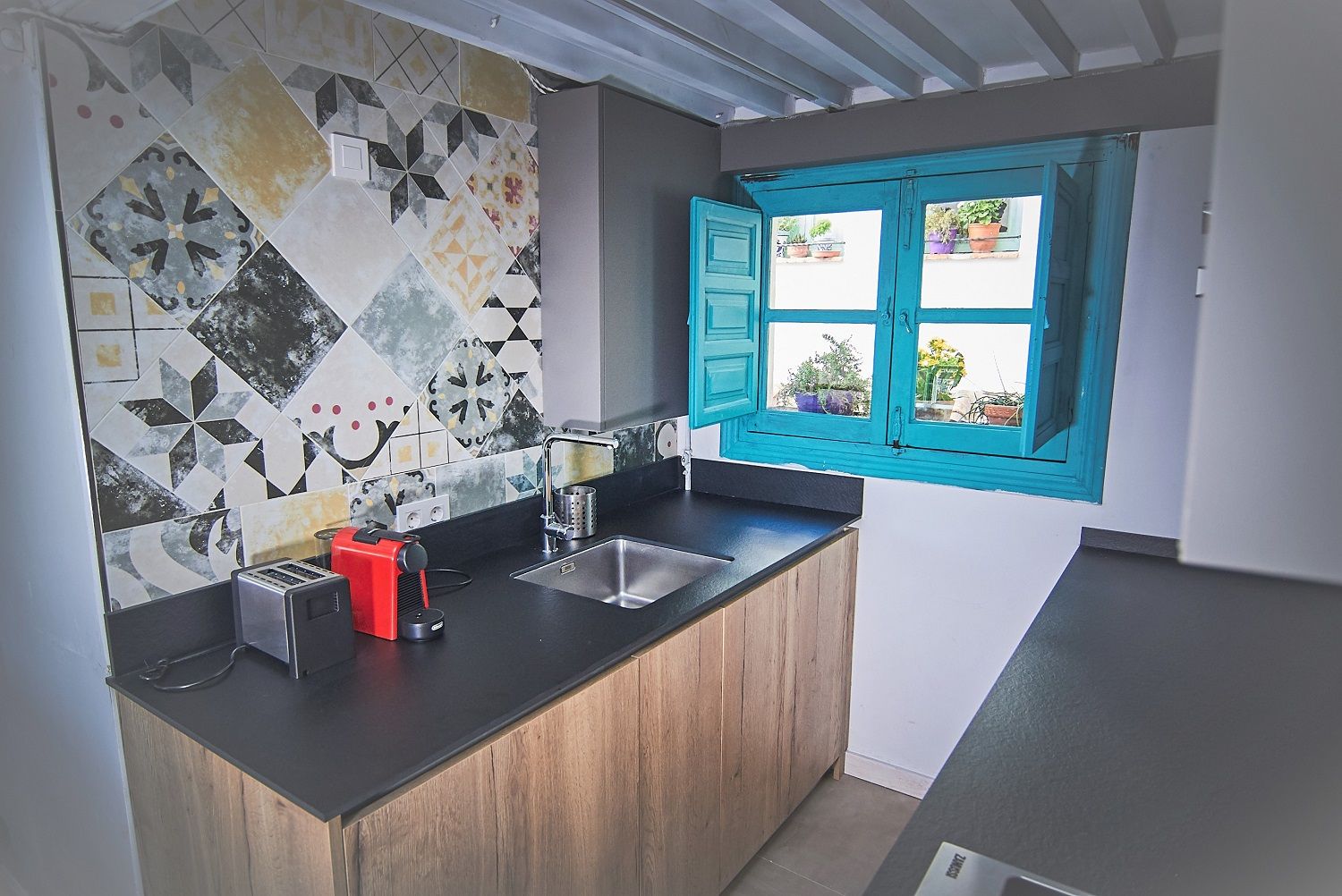 Una cocina con encimera negra, armarios de madera, un electrodoméstico rojo y una ventana con contraventanas azules.