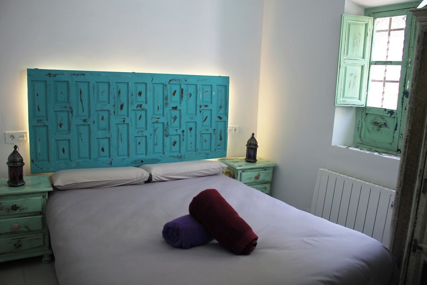 Una cama con sábanas blancas, un cabecero color turquesa y dos toallas enrolladas.