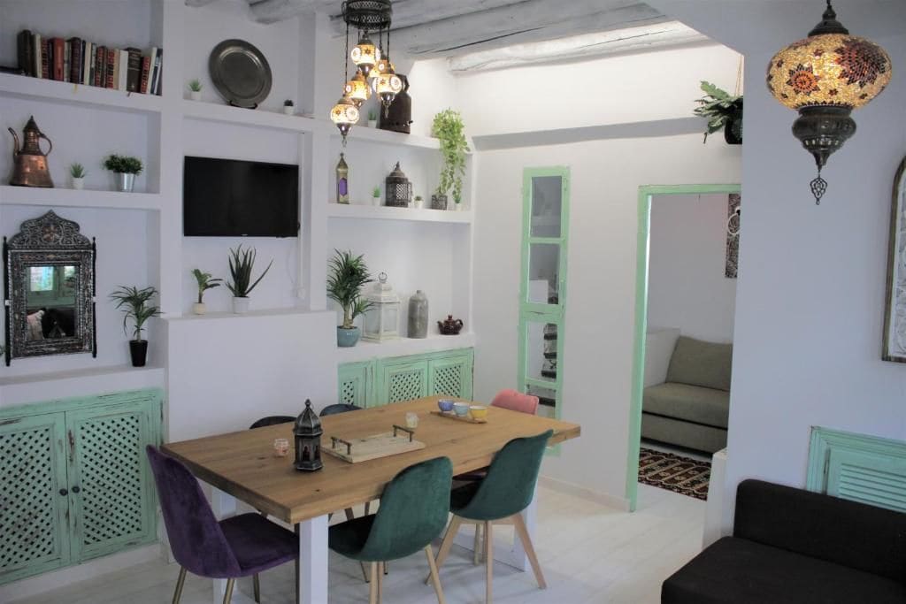 Een eetgedeelte met een houten tafel en stoelen, een flatscreen televisie aan de muur en decoratieve objecten op planken.