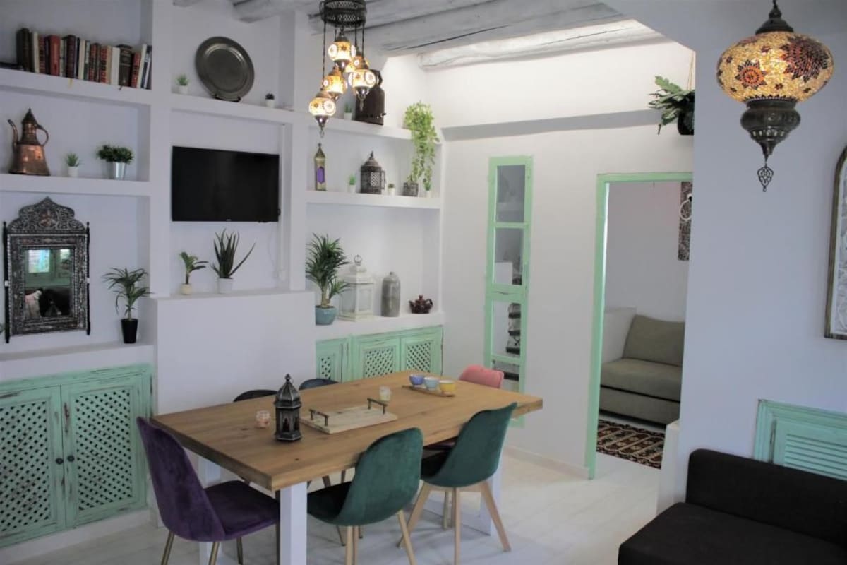 A dining area with a wooden table and chairs, a television mounted on the wall, and shelves with various decorative items.