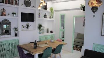 Een eetgedeelte met een houten tafel en stoelen, een flatscreen televisie aan de muur en decoratieve objecten op planken.
