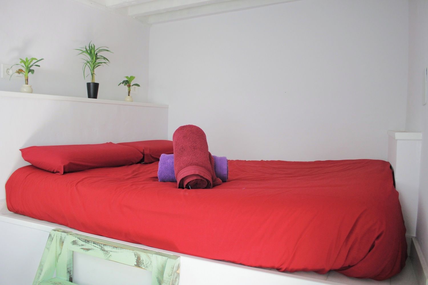 Un lit avec une literie rouge et une serviette enroulée, une petite étagère avec des plantes en pot et un mur blanc.