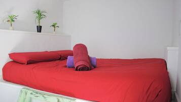 Un lit avec une literie rouge et une serviette enroulée, une petite étagère avec des plantes en pot et un mur blanc.