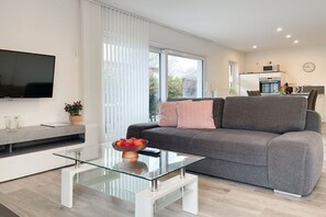Una sala de estar moderna con un sofá gris, una mesa de centro de vidrio con fruta, un televisor de pantalla plana y una zona de cocina al fondo.
